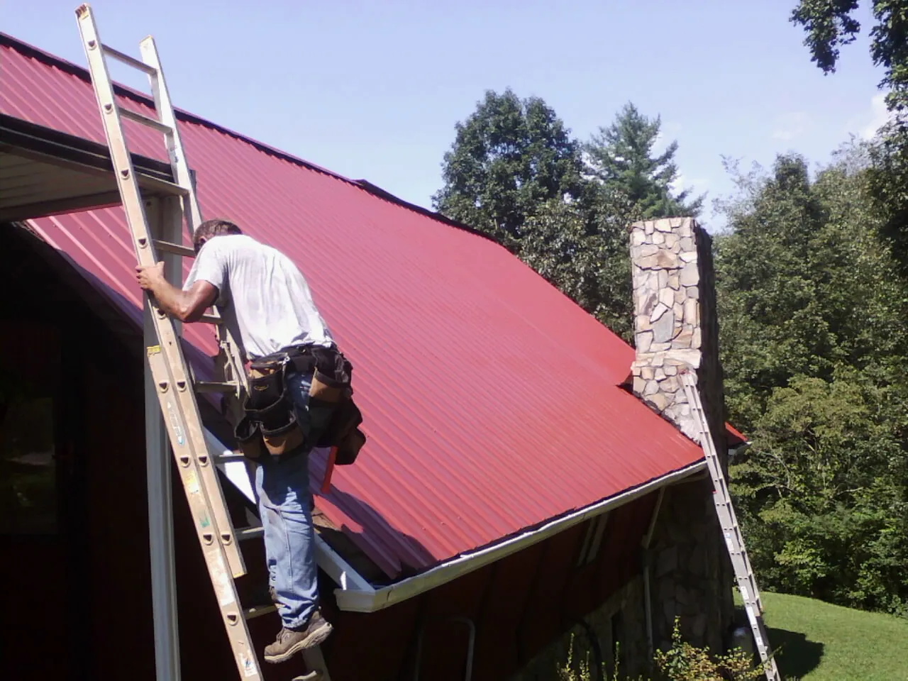 Roofer on a ladder beside a red metal roof and stone chimney — Soffit & Fascia Repair by Roofing Brothers in Peapack